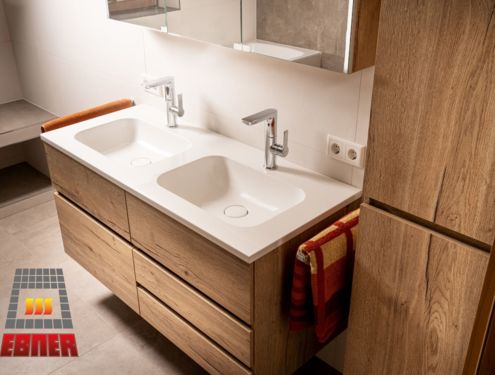 Modern, zeitlos und im Landhausstil. Durch die schlichten Fliesenfarben und den Badezimmermöbeln aus Holz wirkt das Badezimmer gemütlich und freundlich.