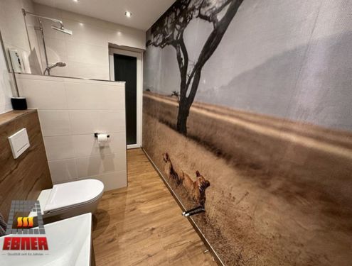 Ein extravagantes Badezimmer. Die Holzoptikfliesen und weißen Fliesen sind zwar schlichter gehalten, doch durch die ganzflächige Fototapete wird das Badezimmer zur Savanne.